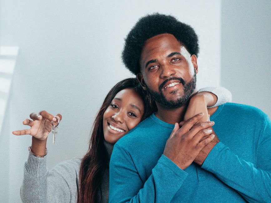 A smiling couple stands proudly in front of their beautiful suburban home, holding up a set of keys to celebrate a successful and profitable home sale.