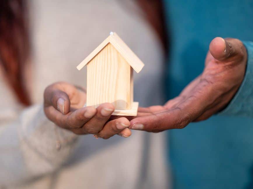 Pair of hands carefully cradling a small, white model of a house against a bright, clean background, symbolizing the prote...