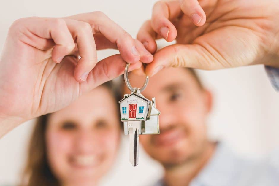 A smiling couple proudly holding the keys to their new home, which has a 'Sold' sign in the front yard, symbolizing the financial savings and success of homeownership.