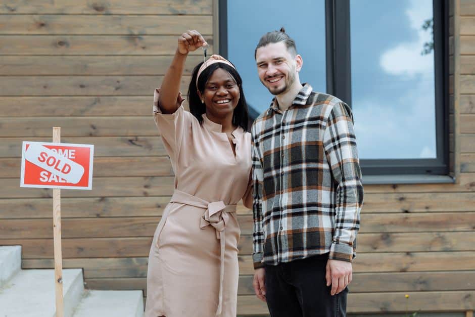A smiling couple stands proudly in front of their beautiful suburban home with a 'Sold' sign in the front yard, symbolizing a successful home sale and preserved equity.
