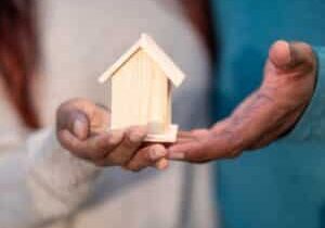 Pair of hands carefully cradling a small, white model of a house against a bright, clean background, symbolizing the prote...